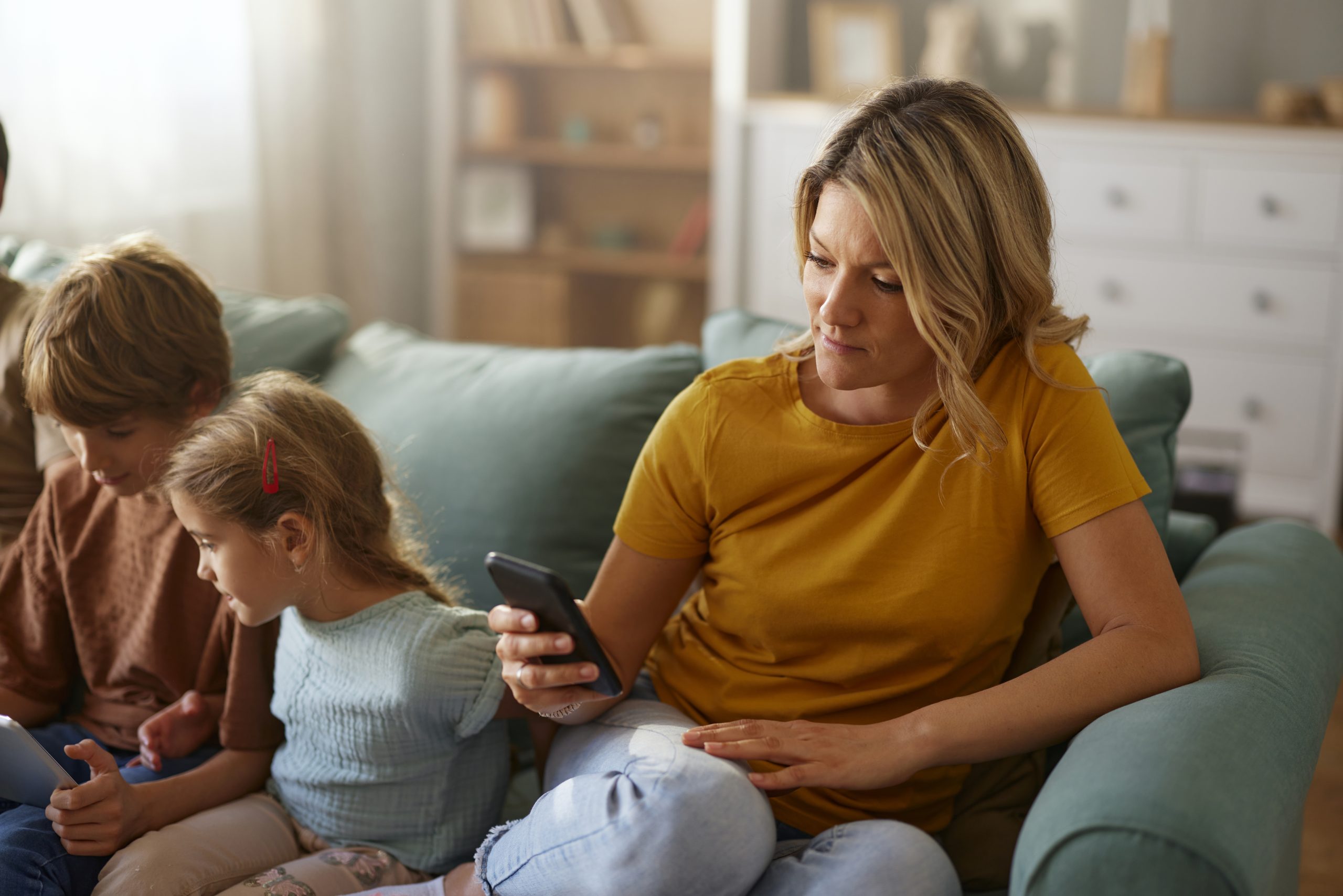 Carefree mother text messaging on smart phone while relaxing next to her kids on sofa in the living room.