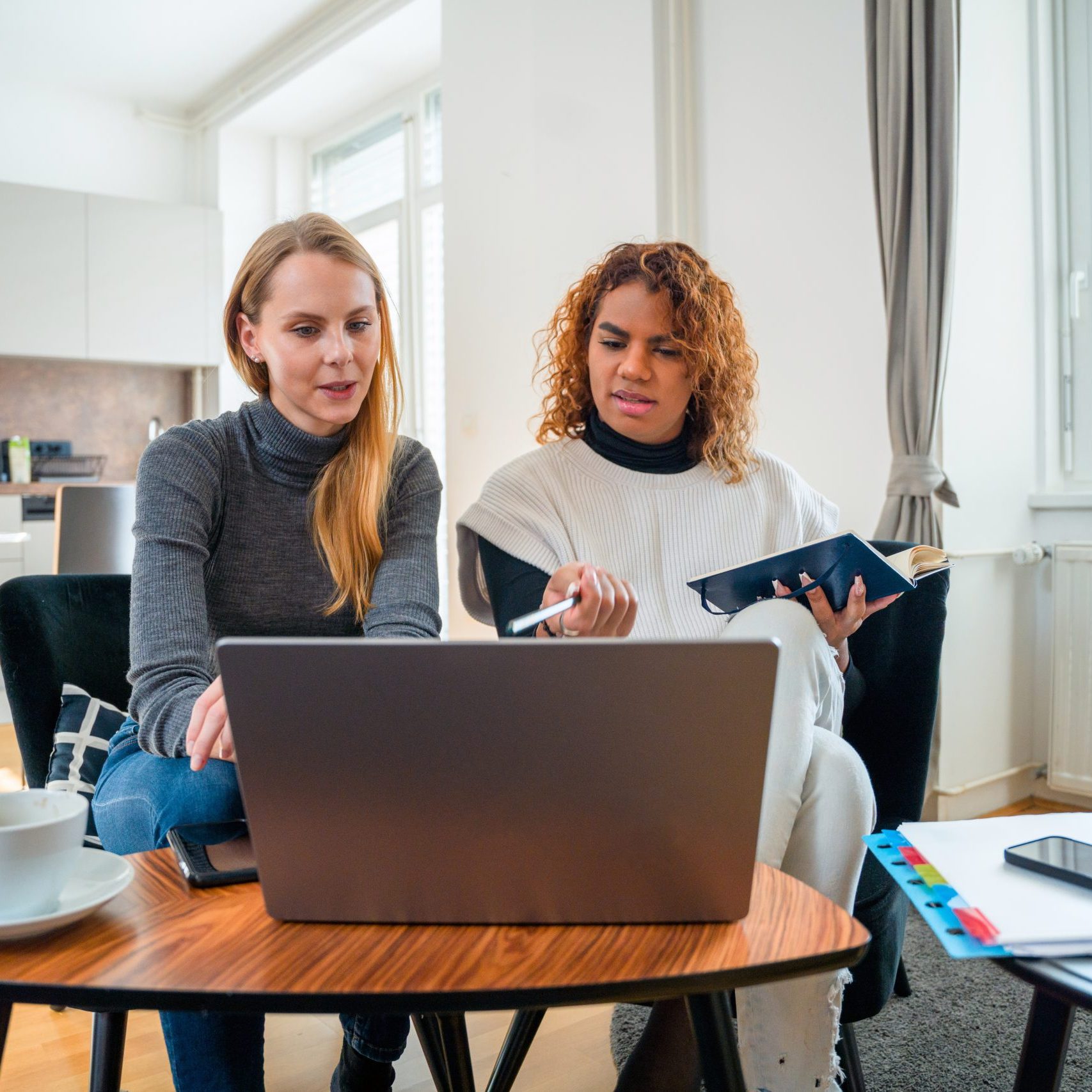 A female and one non-binary gender person are sitting in the living room at home and using laptop together. They are spending time together.