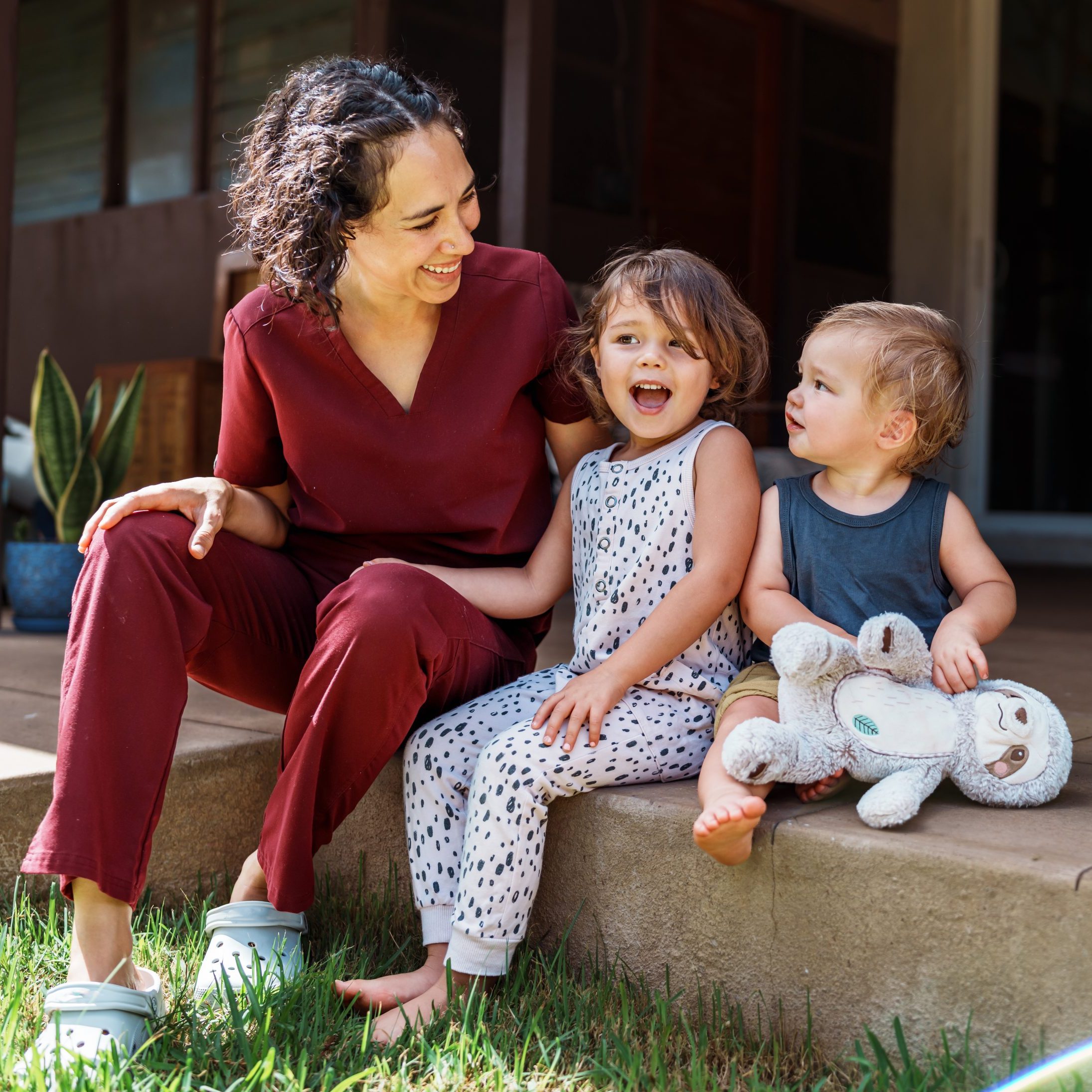 A female nurse of Hawaiian and Chinese descent sits outside on the back patio of her home and playfully interacts with her cheerful and cute Eurasian son and daughter after returning home from work.