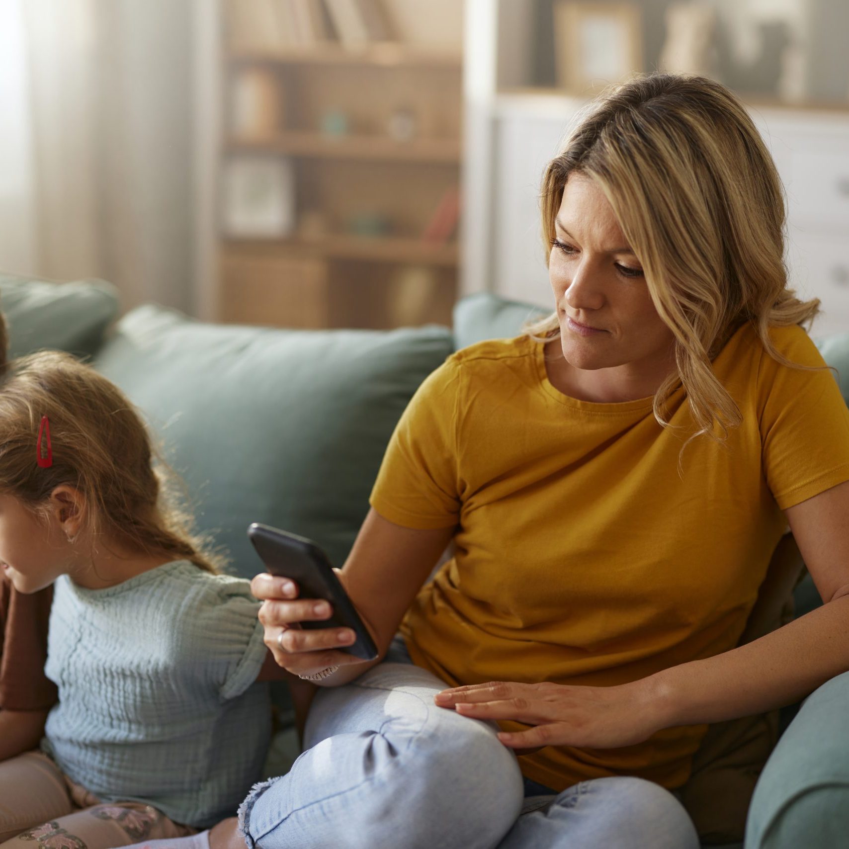 Carefree mother text messaging on smart phone while relaxing next to her kids on sofa in the living room.
