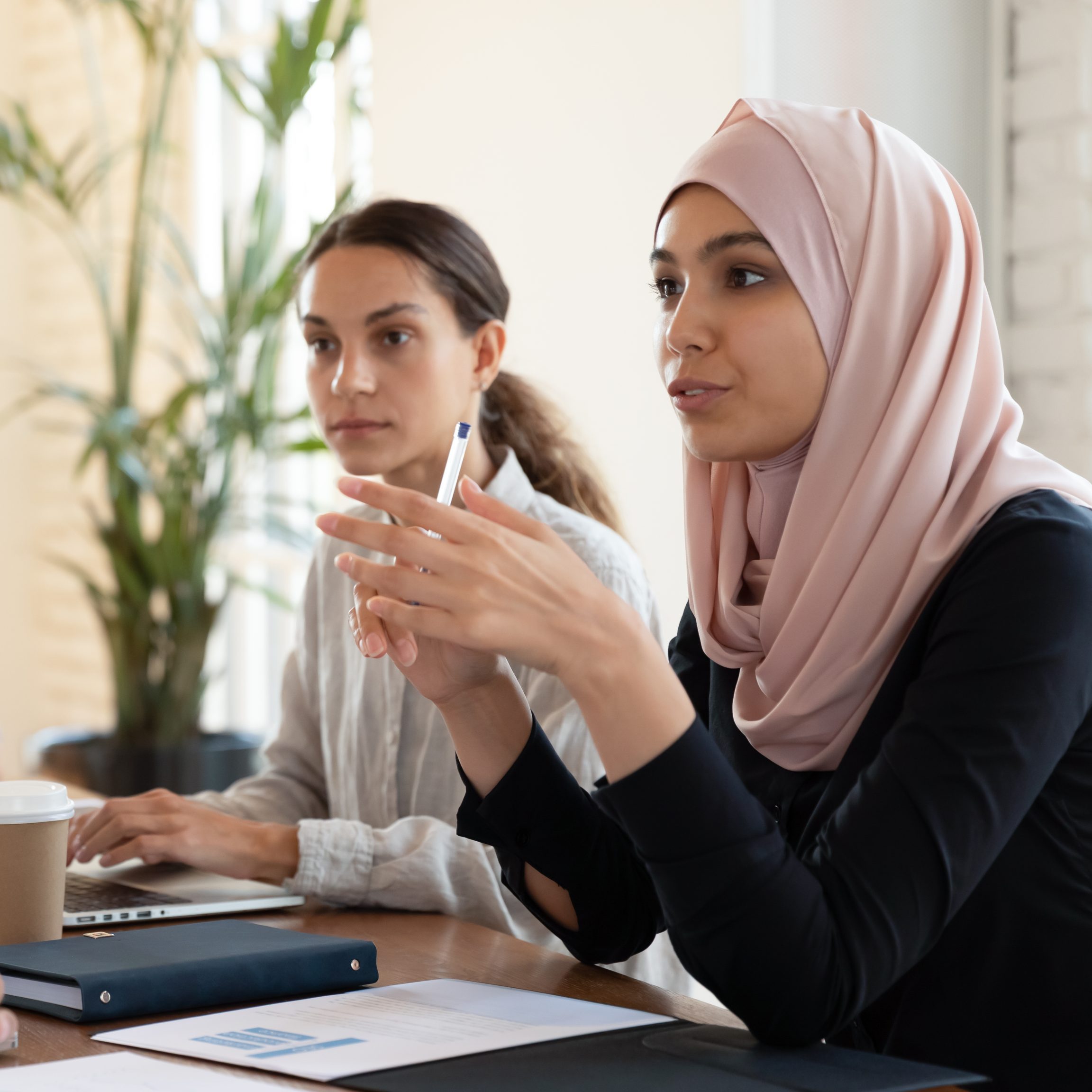 Multicultural businesspeople negotiating gathered together at office group meeting led by businesswoman middle eastern ethnicity leader in hijab, talking expressing opinion speak with european partner