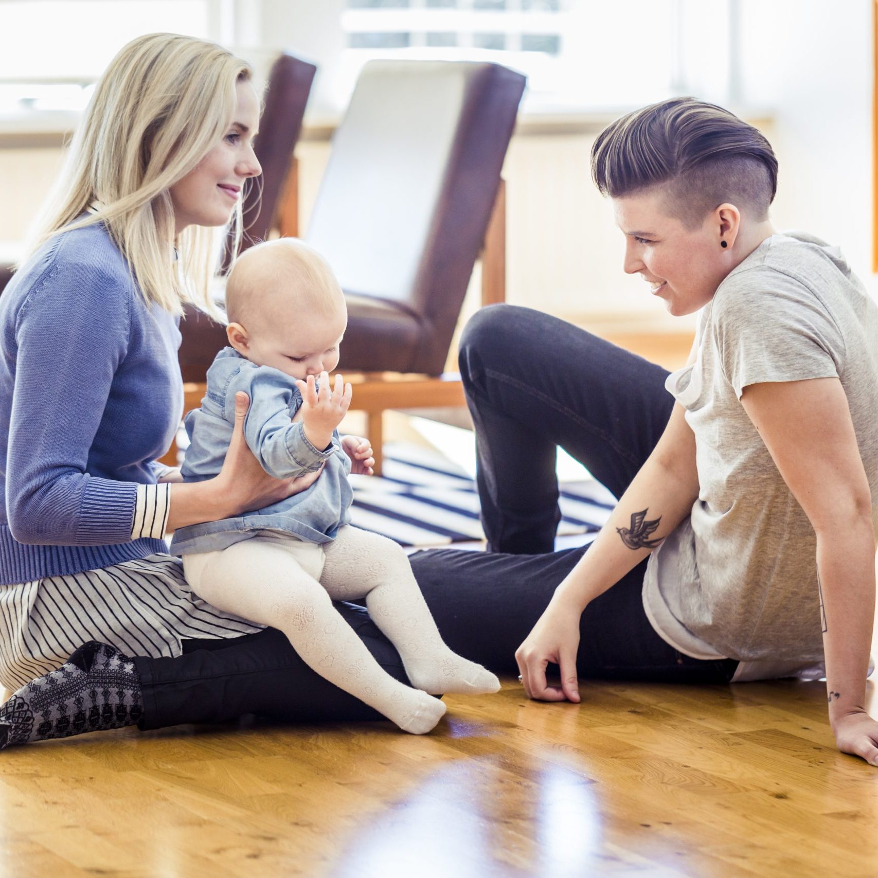 Loving young lesbian couple with baby in living room. Homosexual couple with toddler sitting on hardwood floor. Happy family of three at home.